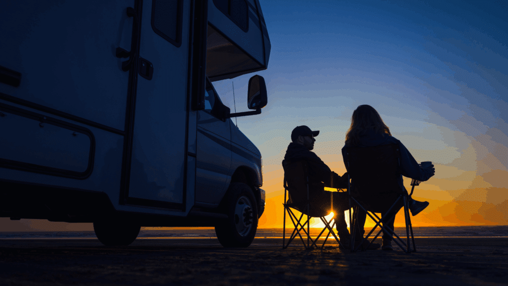Couple relaxing beside their RV while watching the sunset at a beachfront campground.