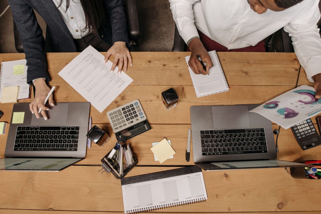 Two people reviewing charts and planning documents at a desk with laptops, calculators, and a calendar