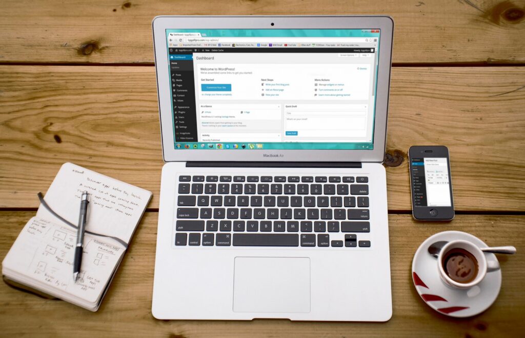 MacBook Air open to a WordPress dashboard beside a handwritten notebook, smartphone, and coffee on a wooden desk