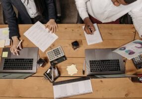 Two people reviewing charts and planning documents at a desk with laptops, calculators, and a calendar