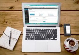 MacBook Air open to a WordPress dashboard beside a handwritten notebook, smartphone, and coffee on a wooden desk